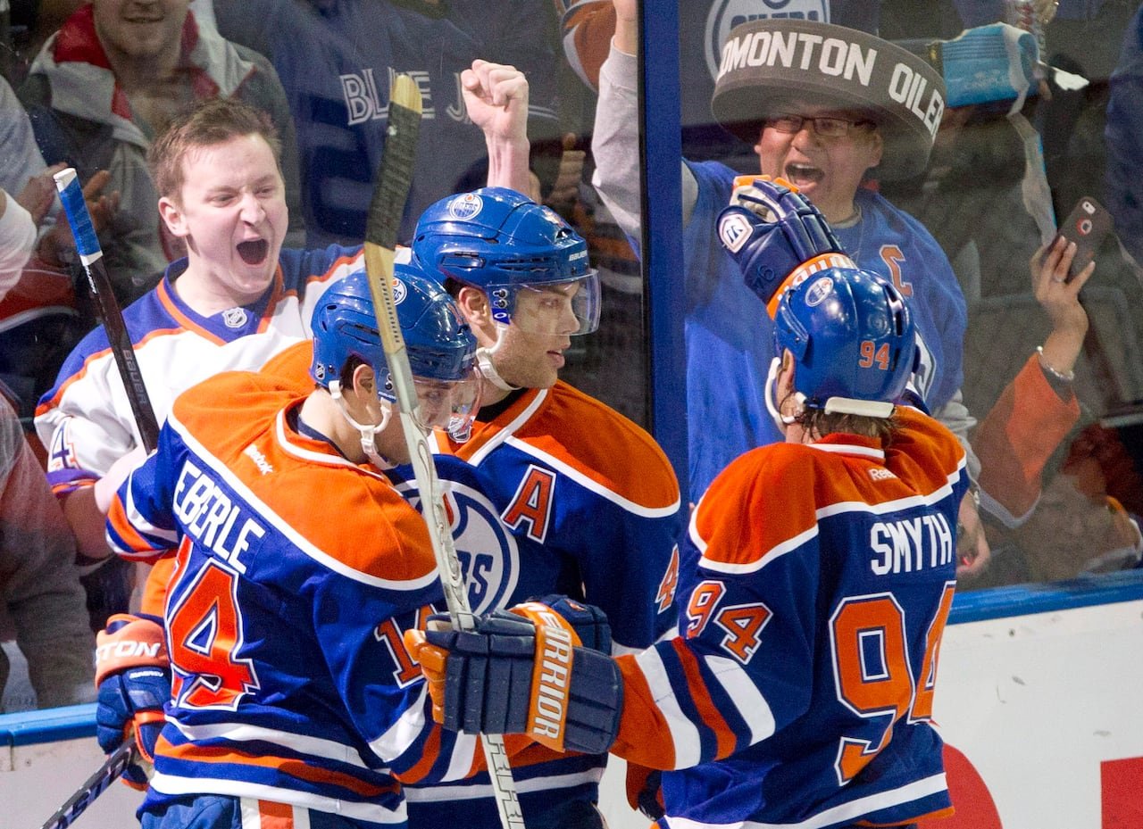 Three hockey players in orange-and-blue jerseys embrace and celebrate while fans on the other side of the arena glass cheer enthusiastically.