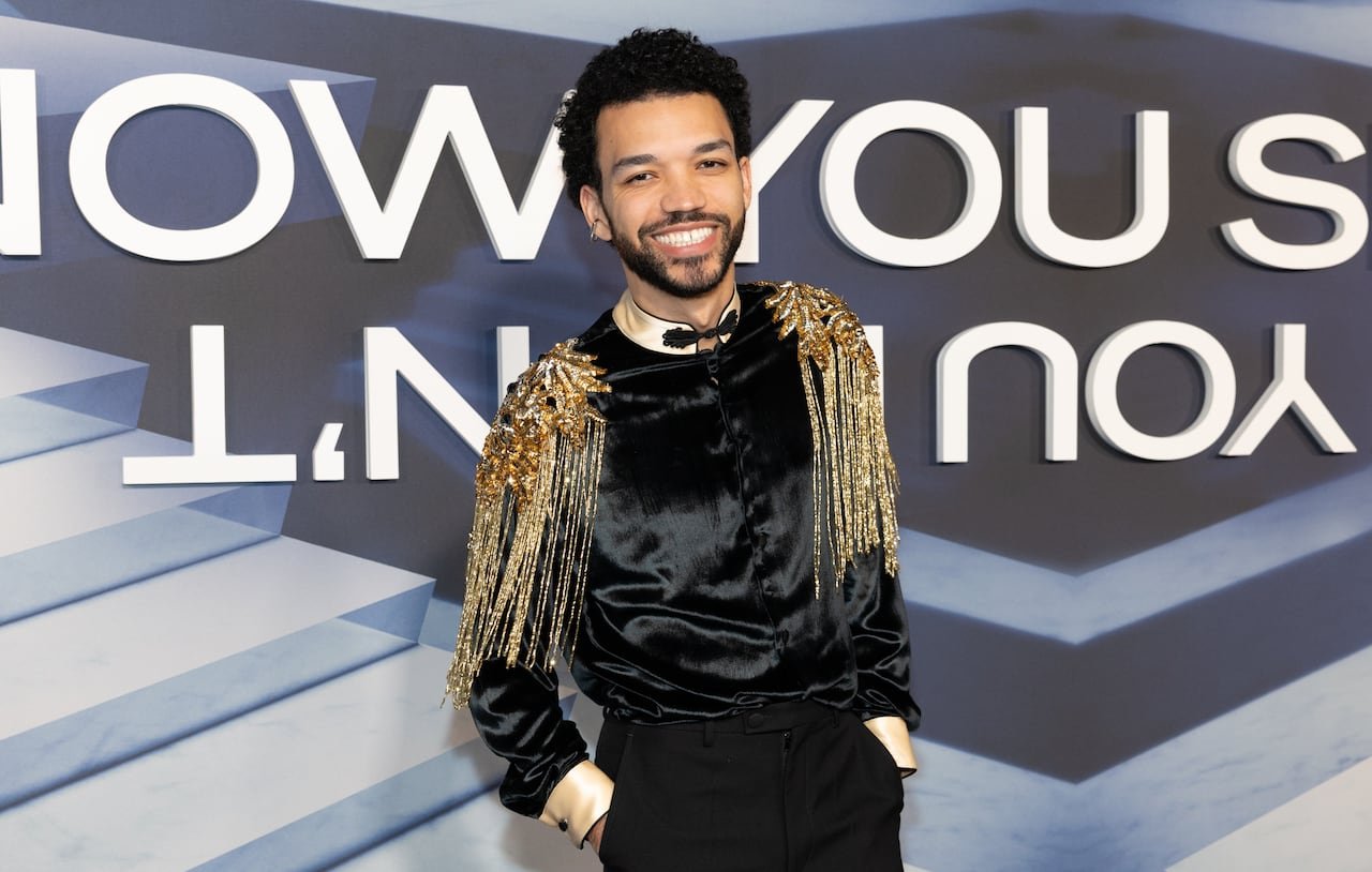 A man poses, smiling, in front of a blue-grey backdrop with white letters.